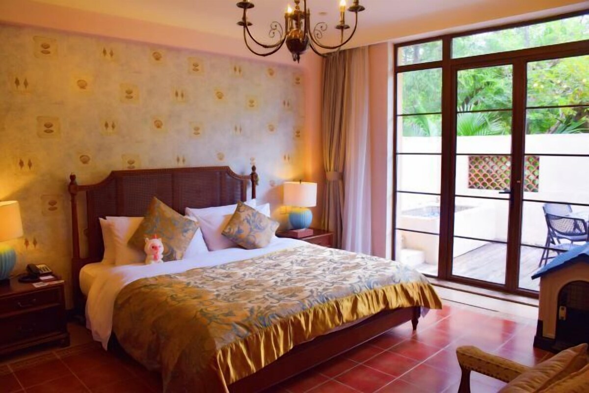 A hotel room with a large bed, a chandelier, a nightstand with a lamp, and a view of greenery through the window.