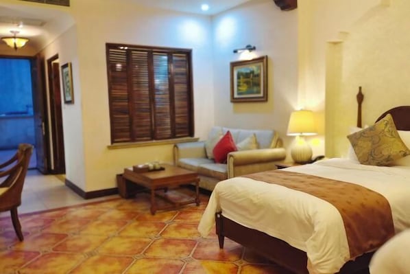 A hotel room with a bed, a sofa, a small table, a window with shutters, and a wall-mounted lamp.