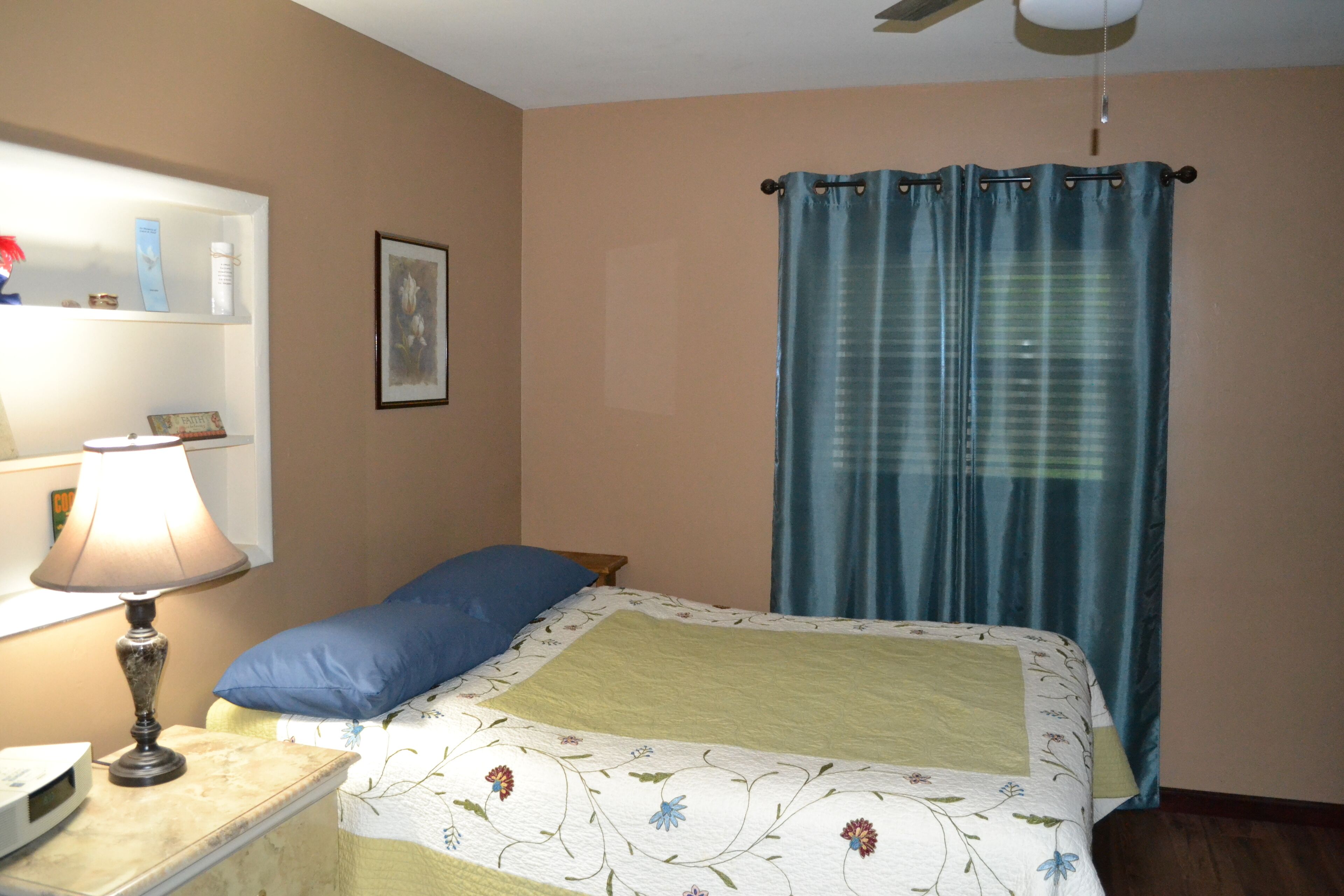 Un dormitorio con cama, mesita de noche con lámpara, ventilador de techo, ventana con cortinas y estante en la pared con objetos.