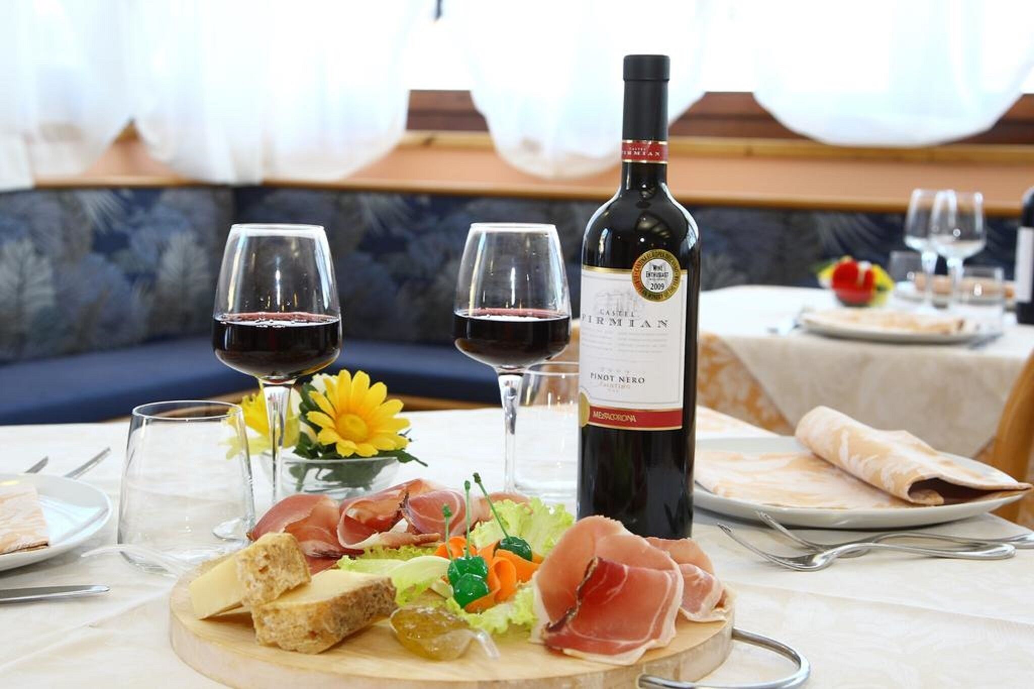 A dining table set with a bottle of Pinot Nero, two glasses of red wine, a variety of cheeses, prosciutto, and a small salad.