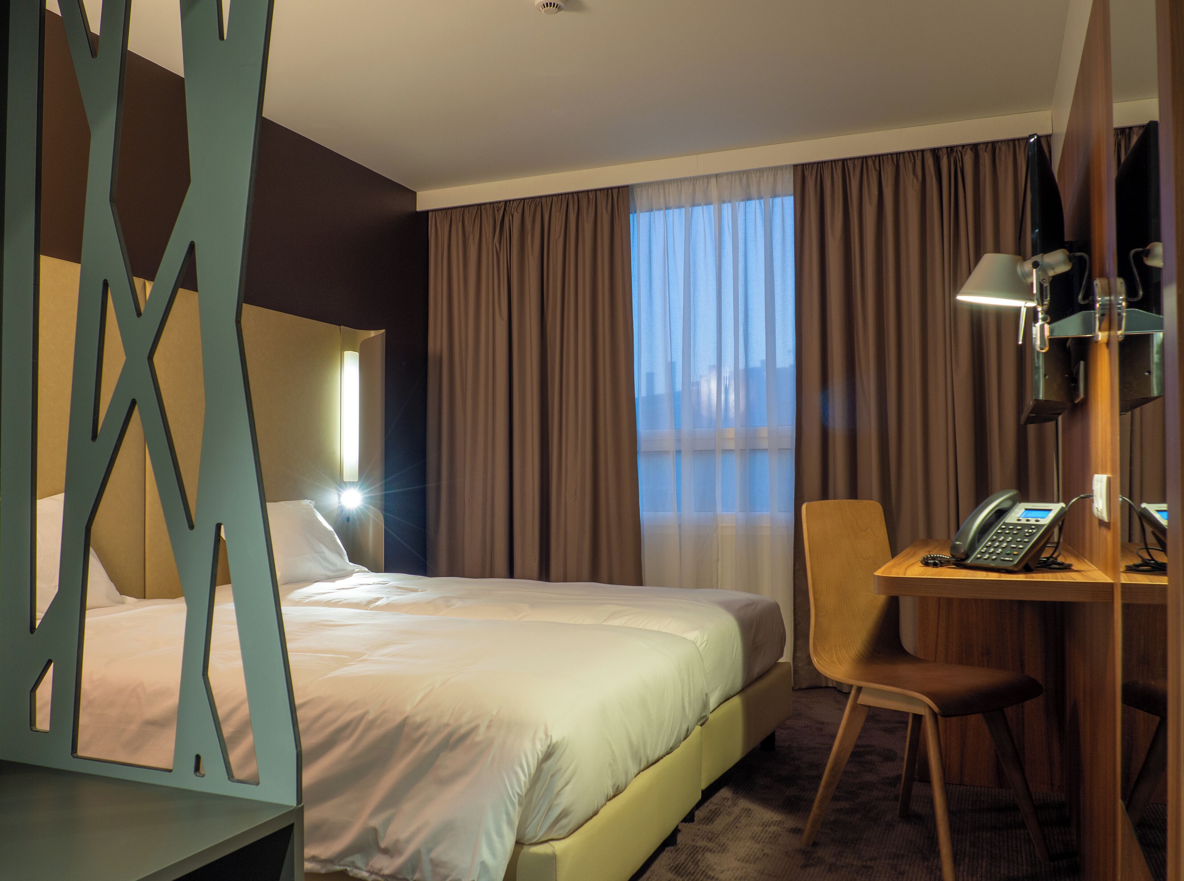 Une chambre d’hôtel avec un lit, un bureau avec un téléphone, une chaise et une grande fenêtre avec des rideaux.