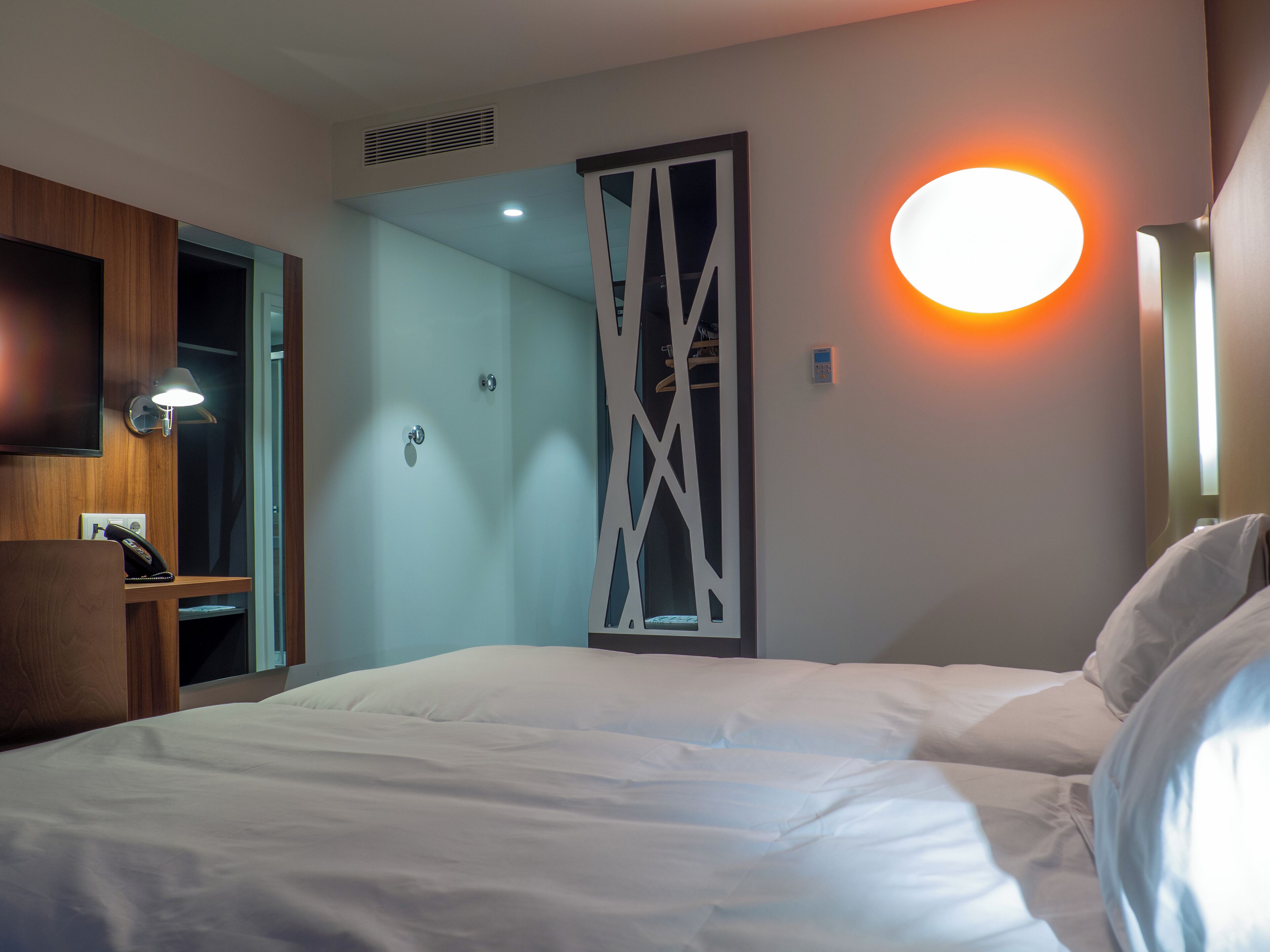 Une chambre d’hôtel avec un lit, un bureau avec une lampe, une télévision et un luminaire fixé au mur.