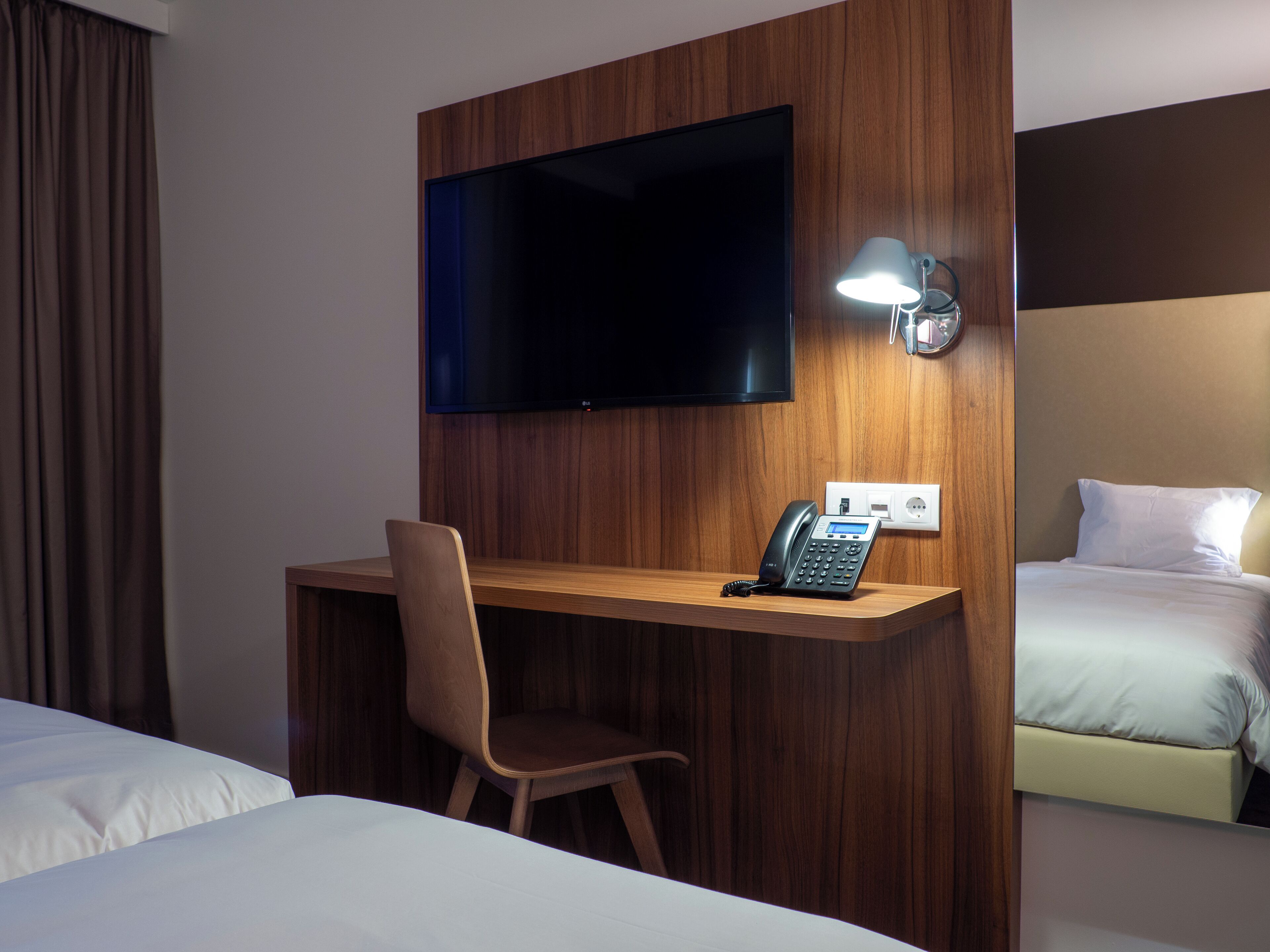 Une chambre d’hôtel avec un bureau en bois, une chaise, un lit, un téléviseur fixé au mur et une table de chevet avec une lampe.