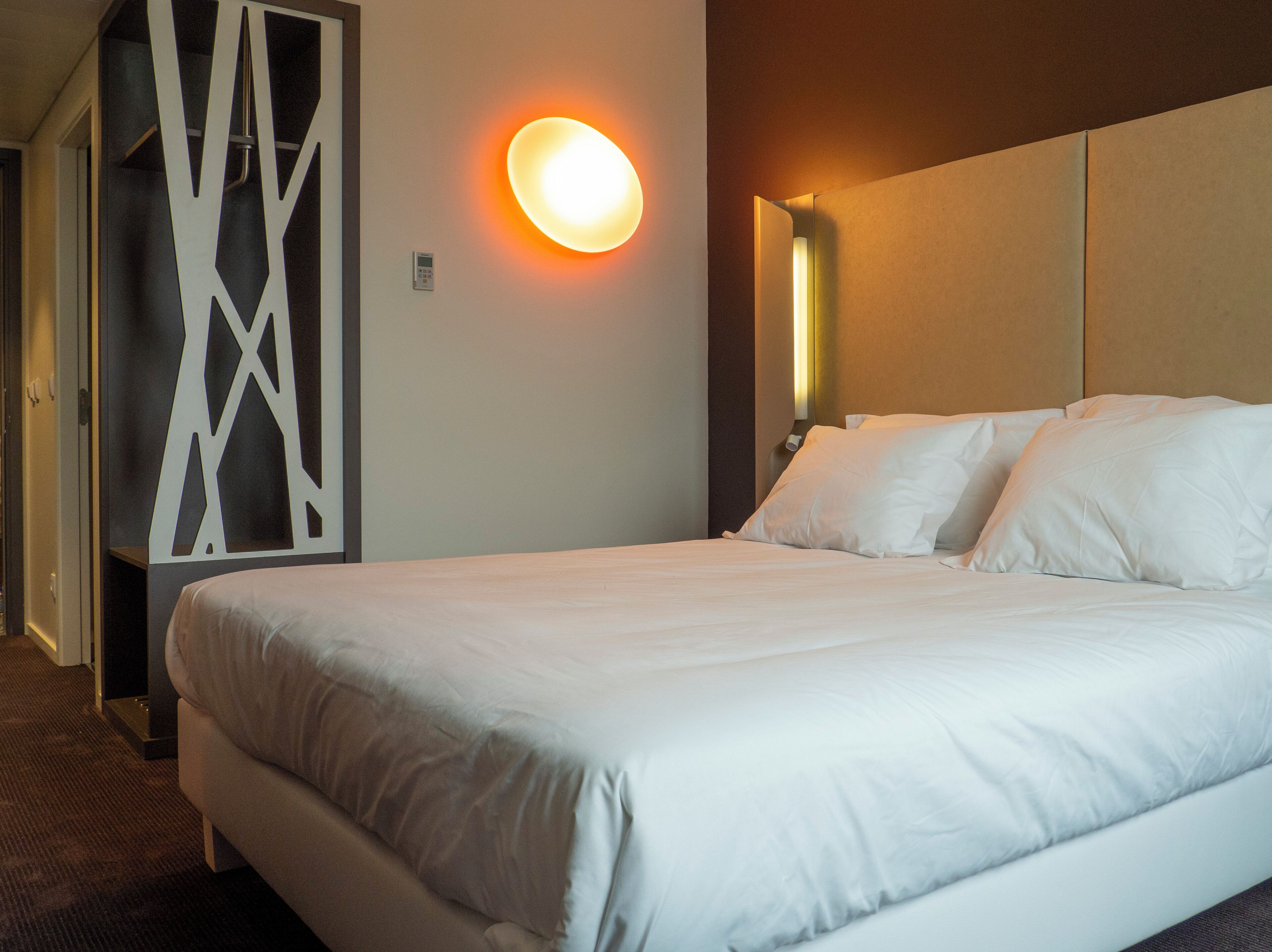 Una habitación de hotel con una cama grande, una lámpara fijada a la pared y un panel decorativo.