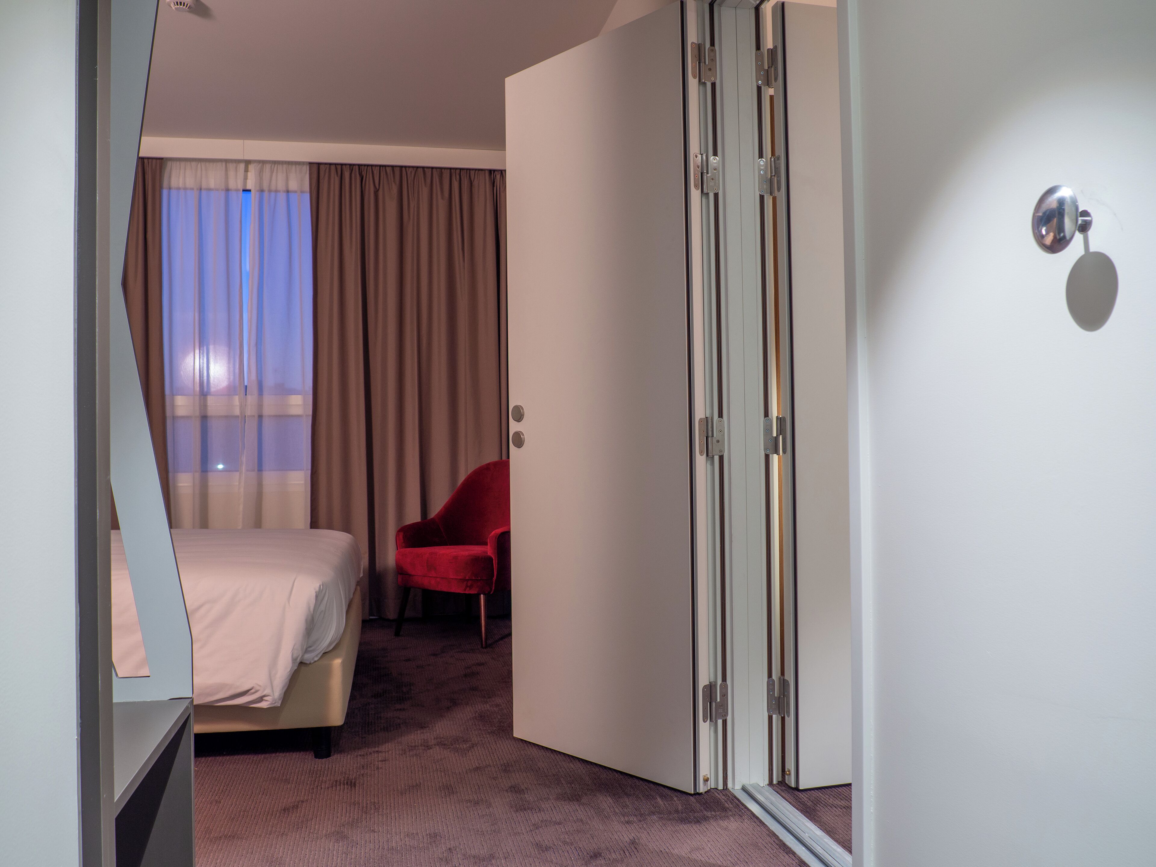 A hotel room with a bed, a red chair, a sliding door, and a view of the outdoors through the window.