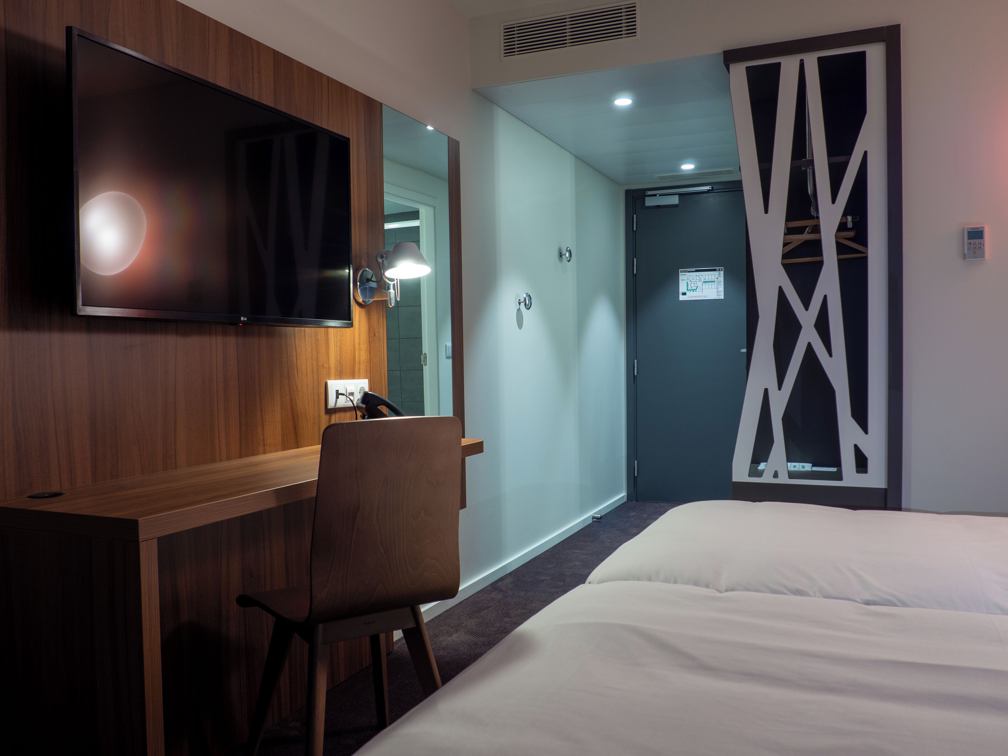 Une chambre d’hôtel avec un bureau en bois, une chaise, un lit, un téléviseur fixé au mur et une porte ornée d’un panneau décoratif.
