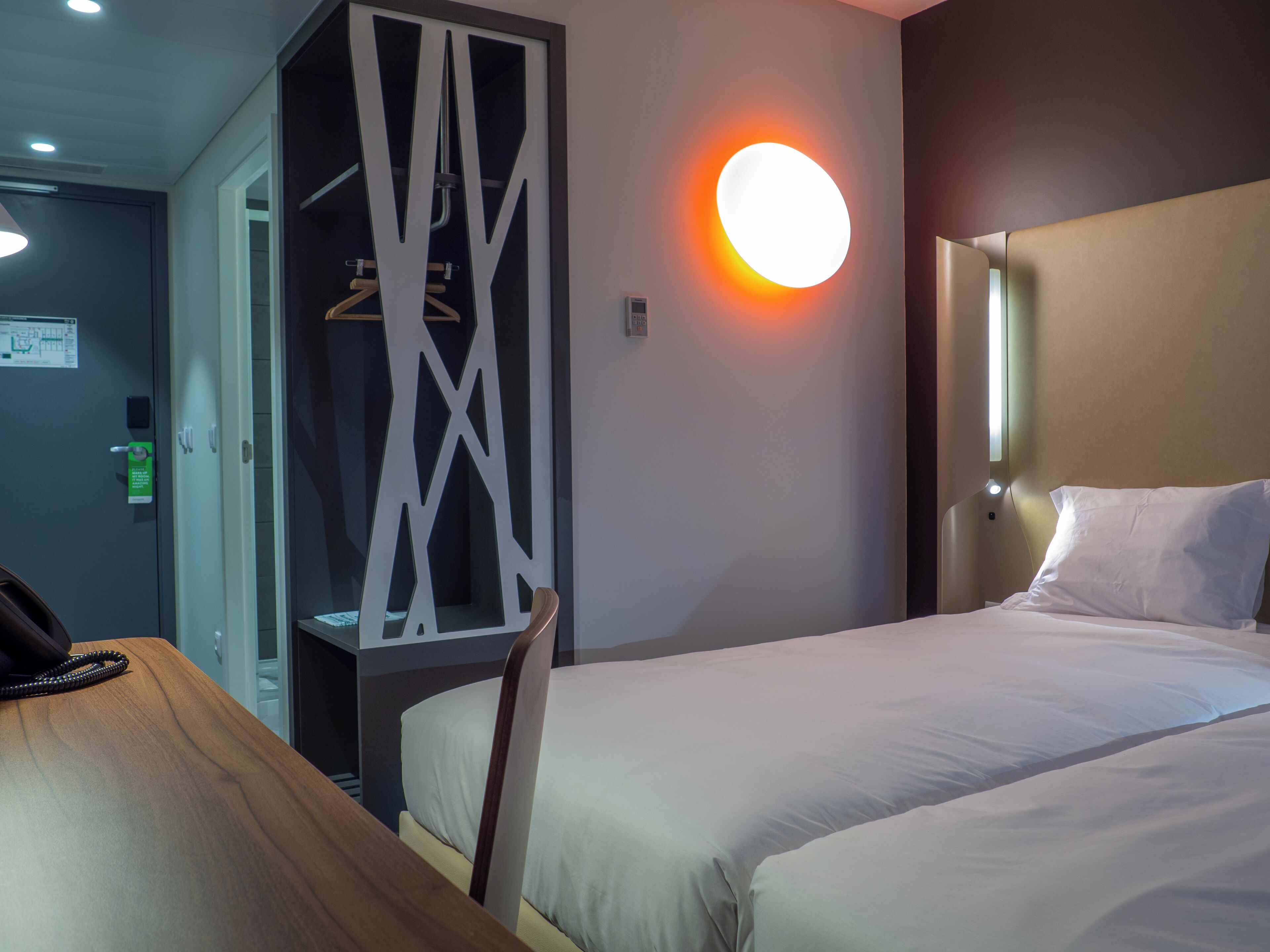 A hotel room with two beds, a wooden desk, a wardrobe, and a wall-mounted light.