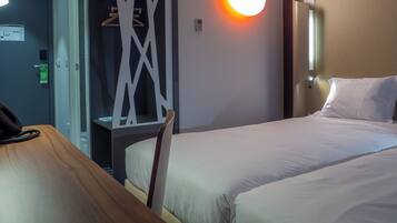 Une chambre d’hôtel avec deux lits, un bureau en bois, une armoire et un lampadaire fixé au mur.