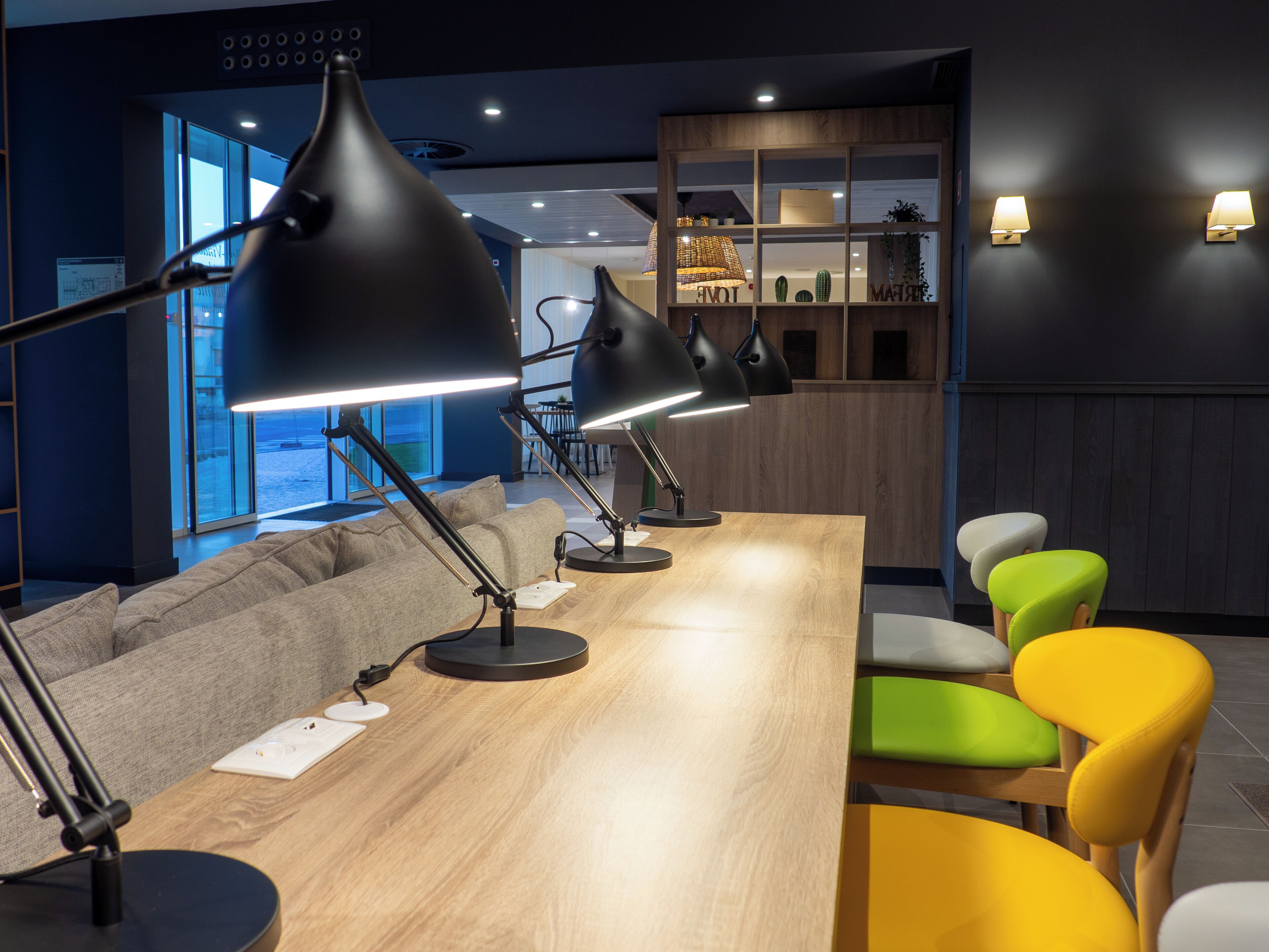 A modern workspace with a long wooden table, multiple black desk lamps, a grey sofa, and colorful chairs.
