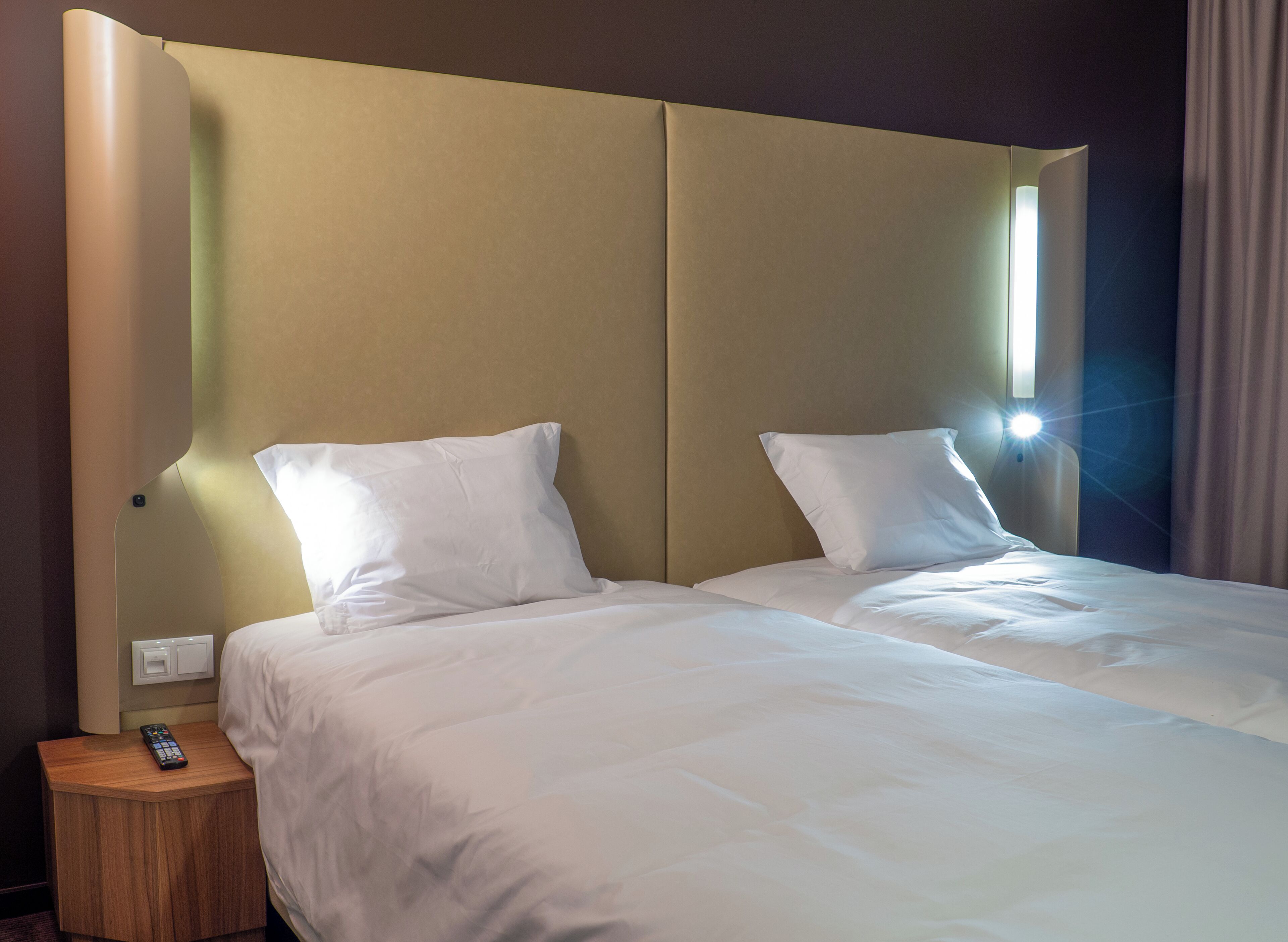 A hotel room with a large bed, two pillows, a nightstand with a remote control, and a wall-mounted light.