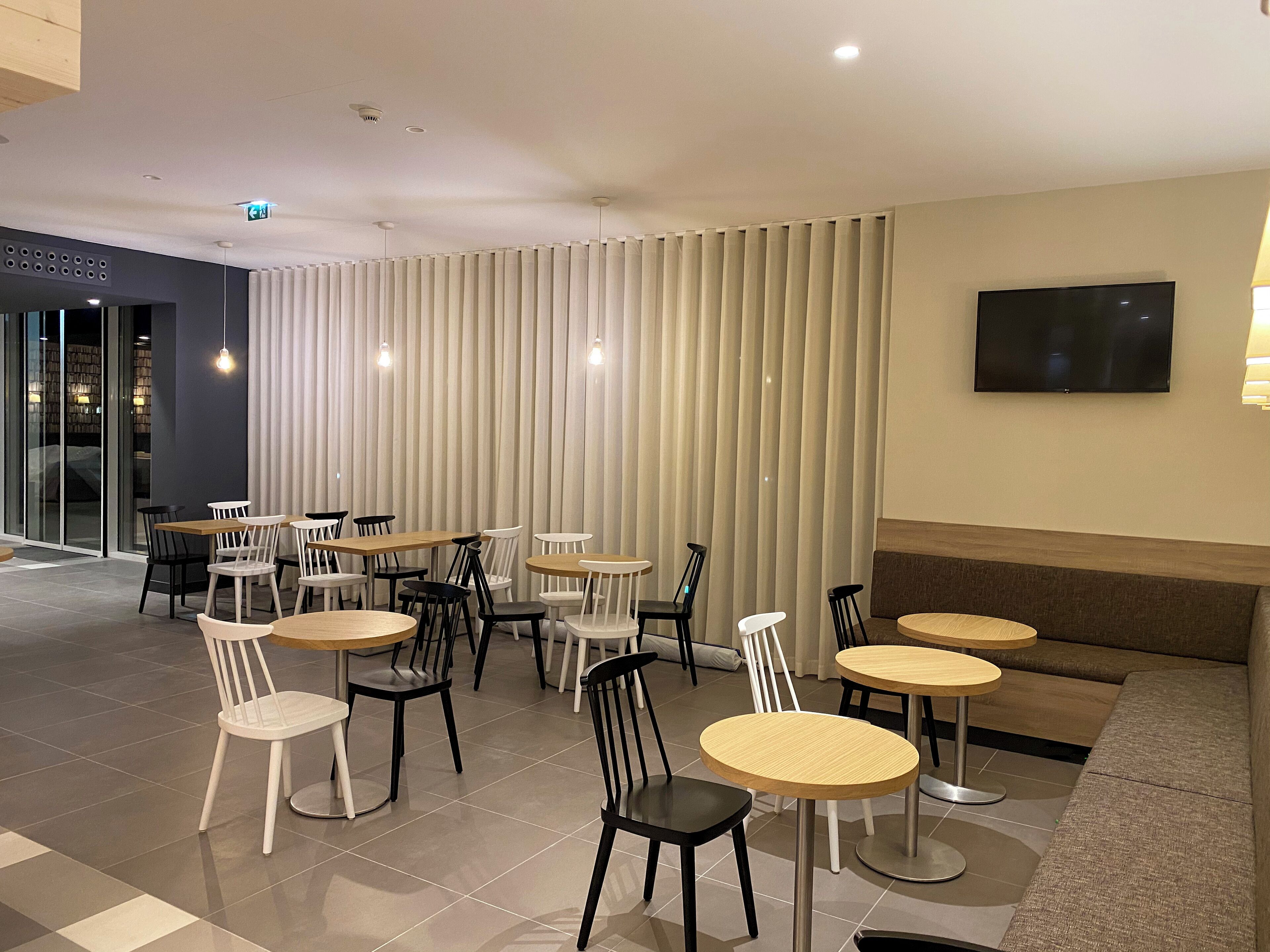 A modern indoor seating area with multiple tables and chairs, a flat-screen TV mounted on the wall, and a large window with blinds.