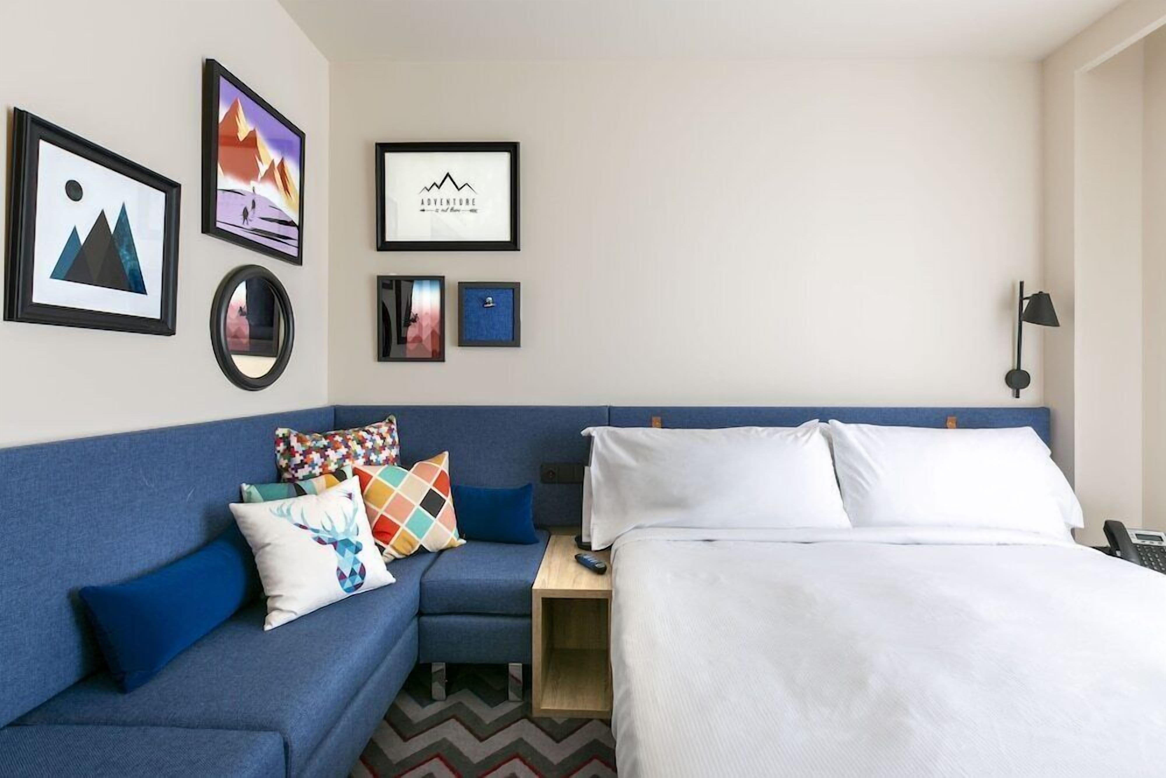 Quarto de hotel com uma cama, um sofá azul, uma mesinha e quadros nas paredes.