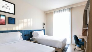 Habitación de hotel con dos camas, un escritorio, una silla, teléfono en la pared y un cuadro enmarcado.