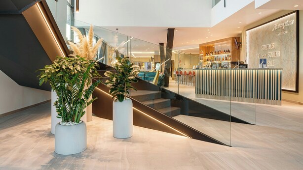 A modern hotel lobby with a glass staircase, a reception desk, and potted plants. - Hampton by Hilton Alcobendas (Alcobendas)