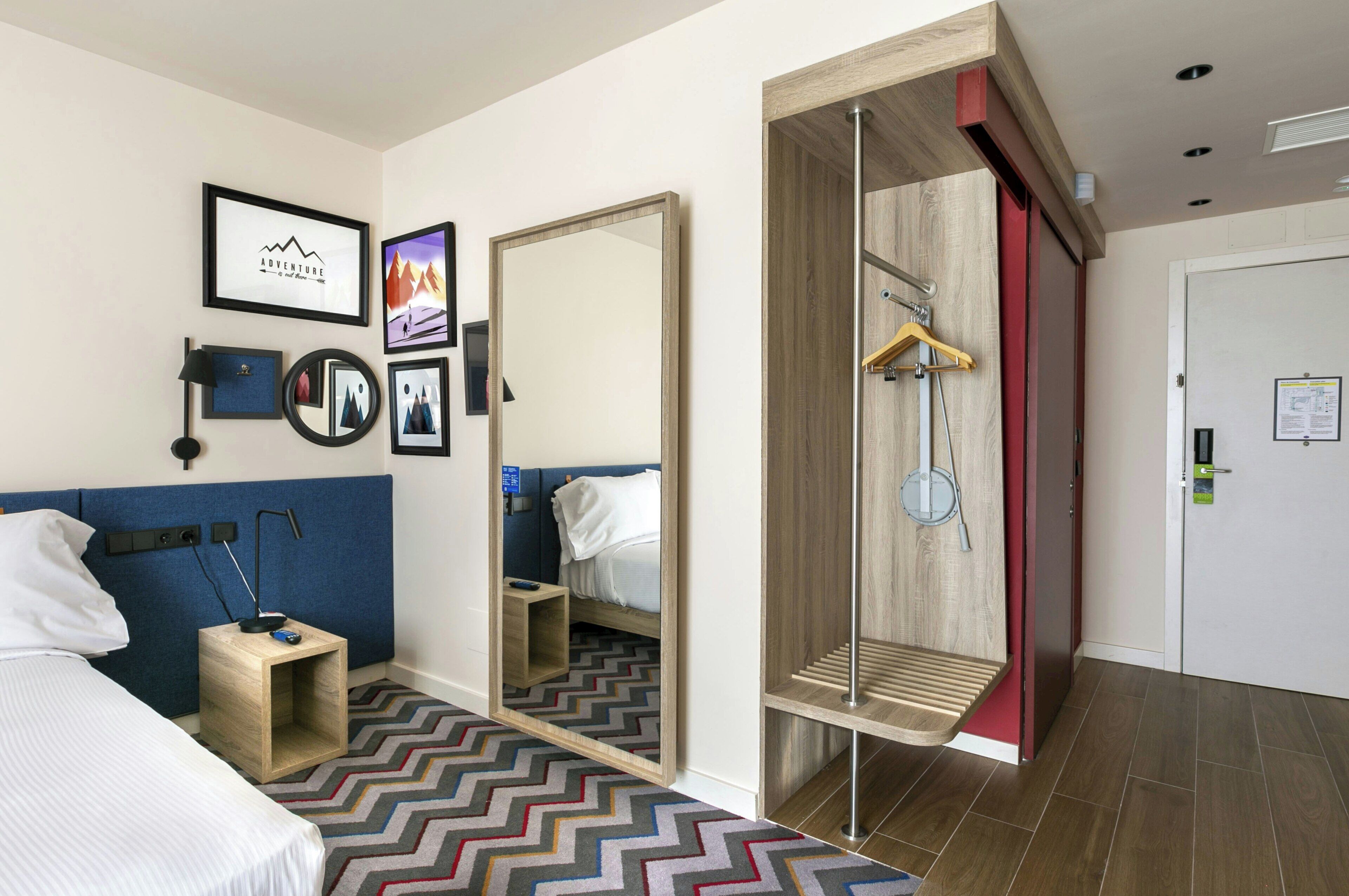Una habitación de hotel con cama, mesita de noche, espejo, armario y una pared decorada con cuadros.