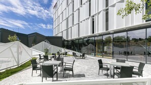 An outdoor seating area with tables and chairs, a glass wall, and a modern building in the background. - Hampton by Hilton Alcobendas (Alcobendas)