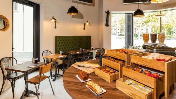 A modern cafe with wooden tables and chairs, a green upholstered bench, and a variety of baked goods displayed in wooden baskets. - Ibis Styles Puteaux Paris La Defense (Puteaux)