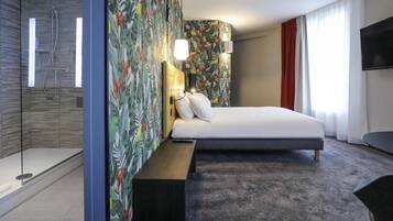 Une chambre d’hôtel équipée d’une douche, d’un lit avec une tête de lit, d’une table de chevet, d’un banc, d’une télévision et d’une fenêtre avec des rideaux.