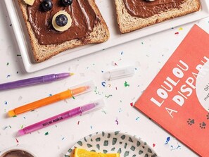 Two slices of toast with chocolate spread and blueberries, colorful markers, a notebook titled 'LOULOU A DISPARU', and a plate with an orange slice. - Ibis Styles Puteaux Paris La Defense (Puteaux)