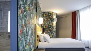 A hotel room with a bed, a bench, a shower, and a wall with a tropical leaf pattern. - Ibis Styles Puteaux Paris La Defense (Puteaux)