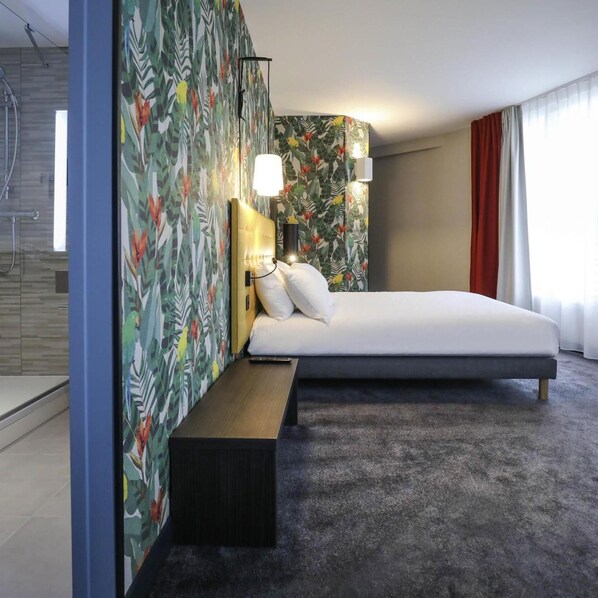A hotel room with a bed, a bench, a shower, and a wall with a tropical leaf pattern. - Ibis Styles Puteaux Paris La Defense (Puteaux)