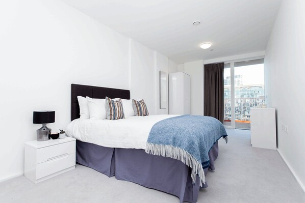 A modern hotel room with a large bed, a bedside table with a lamp, a dresser, and a view of the city through large windows. - Clover Court By Q Apartments (London)