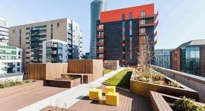 A modern urban building complex with multiple residential and commercial structures, outdoor seating areas, and a rooftop garden.