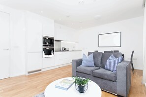 A modern living room with a grey sofa, a coffee table with a book and a plant, and a kitchen area with white cabinets and appliances.