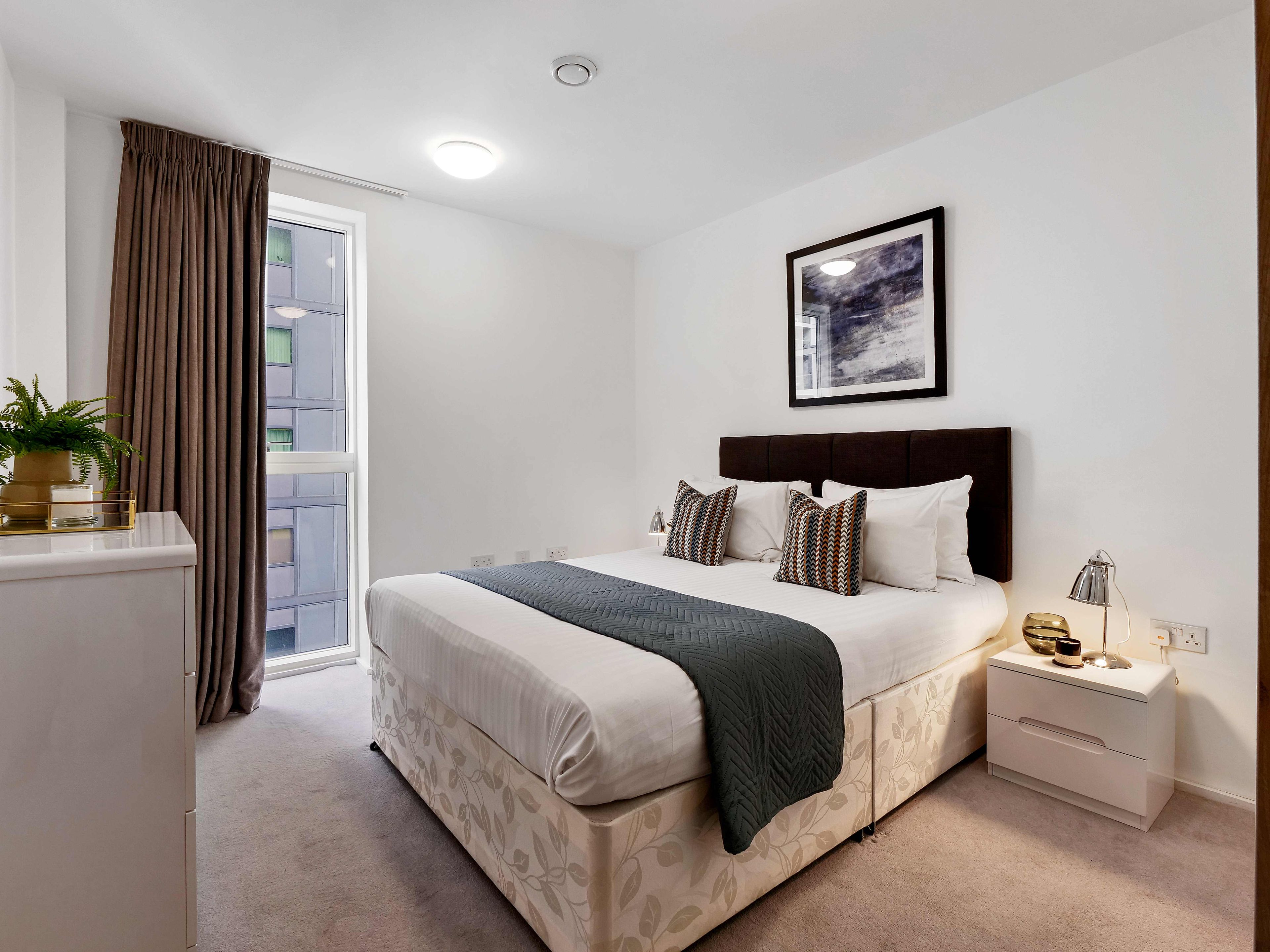 A hotel room with a bed, bedside table, lamp, and a window with curtains.