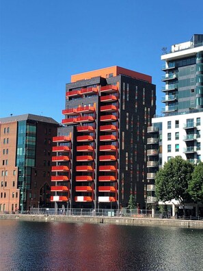 A modern building with multiple balconies, a reflective water body in front, and adjacent buildings.