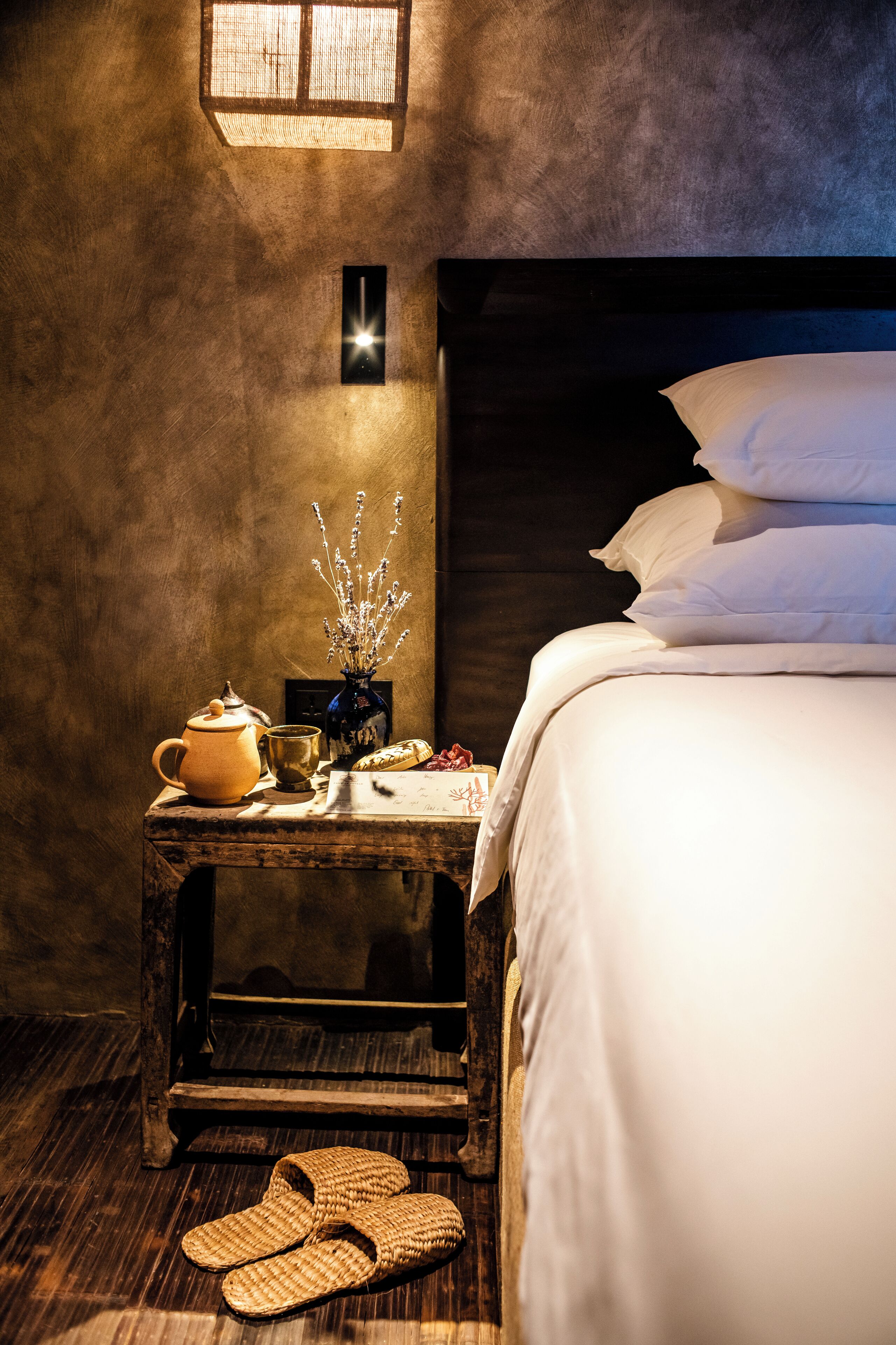 A cozy room with a bed, a nightstand with a lamp, a vase with flowers, a teapot, and slippers.