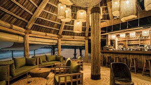 Un bar en la playa con mobiliario de madera, luminarias tejidas y vistas al océano.
