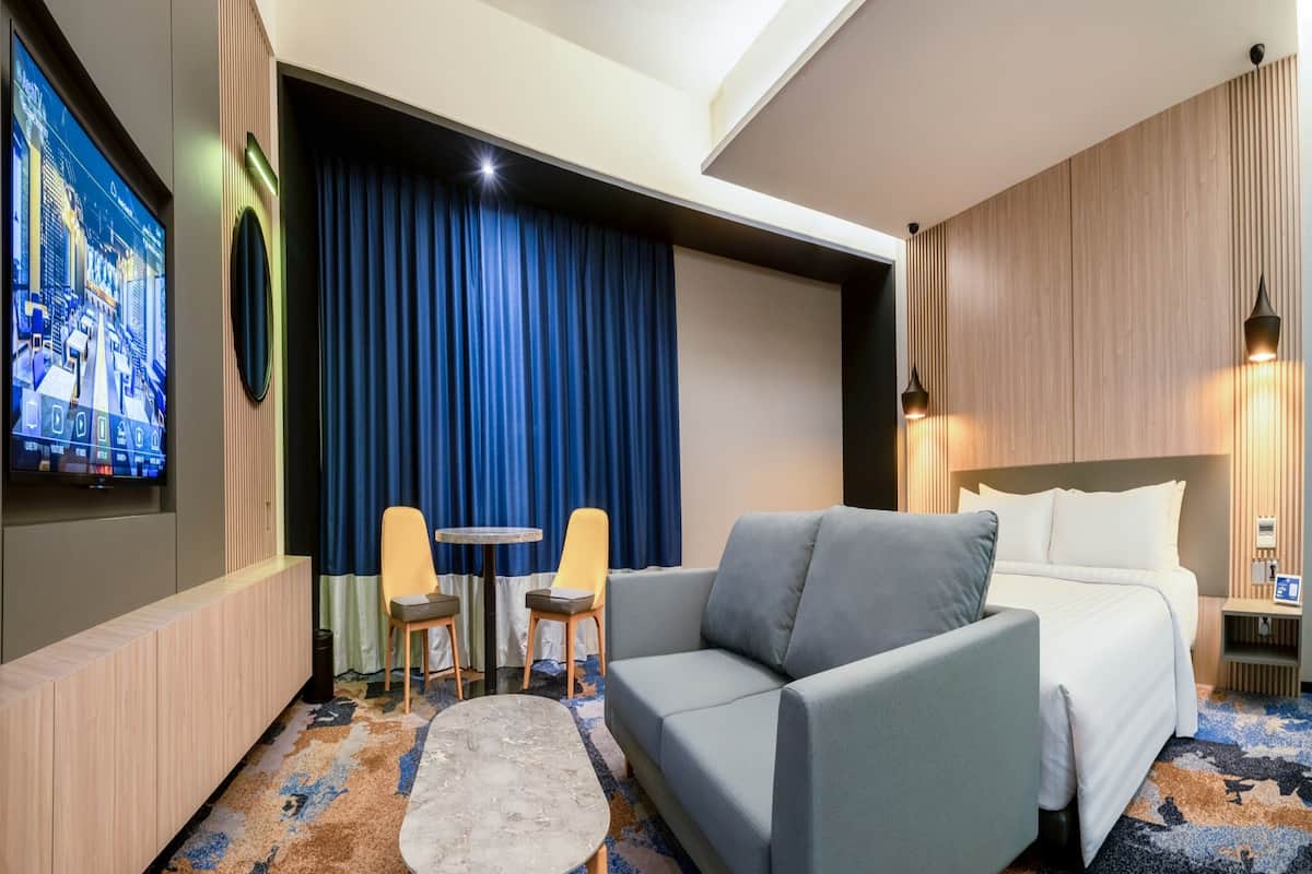 Une chambre d’hôtel avec un canapé gris, une petite table ronde et deux chaises, un lit avec du linge de maison blanc, un grand téléviseur à écran plat fixé au mur et un rideau bleu.
