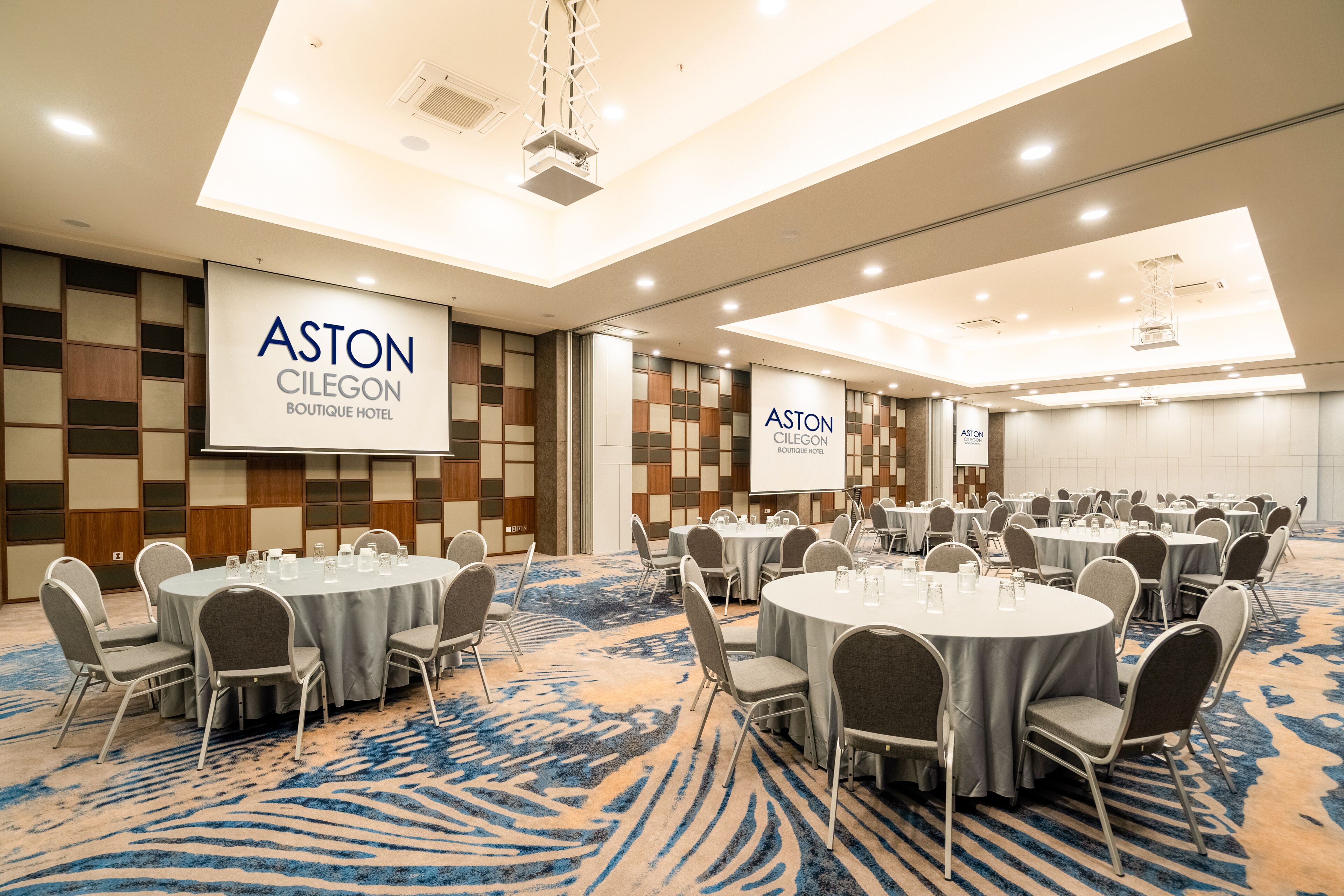 A conference room with round tables and chairs, a blue carpet, and a large screen displaying the hotel name.