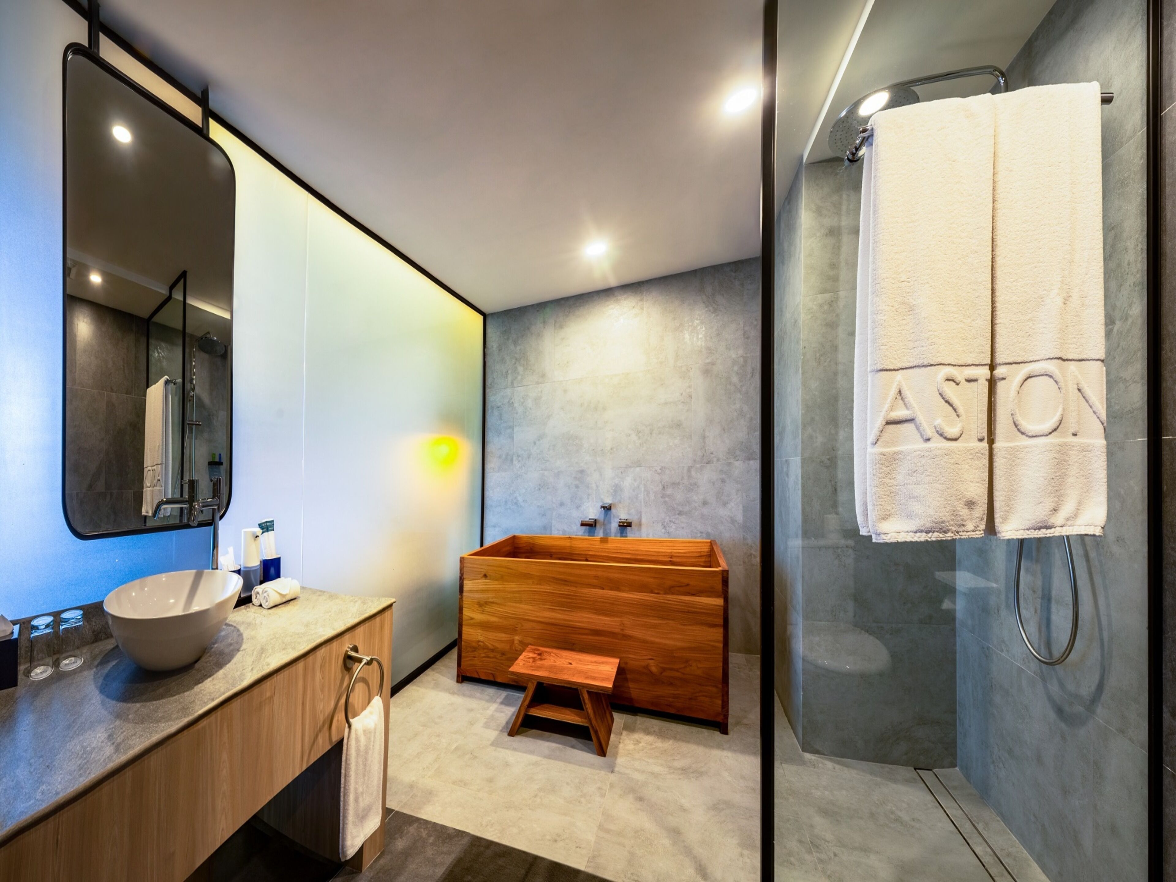 Une salle de bain moderne avec un meuble-lavabo en bois, un grand miroir, une baignoire et un porte-serviettes sur lequel est posée une serviette portant l’inscription « ASTON ».
