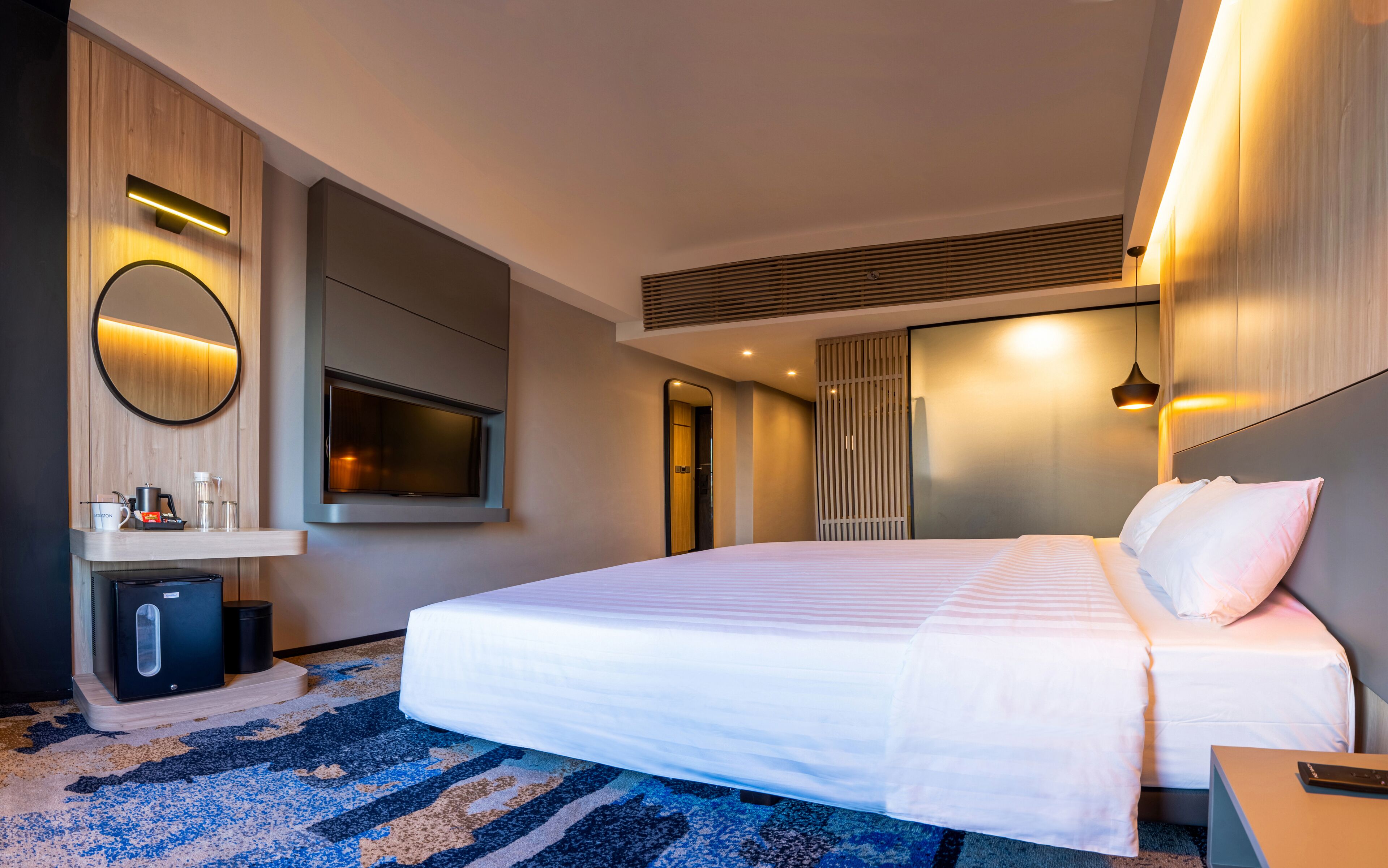 A modern hotel room with a large bed, a flat-screen TV, a bedside table, a mirror, and a built-in heater.