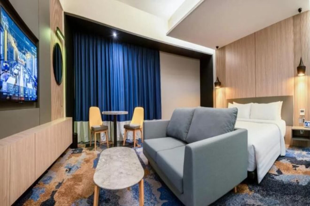 Una habitación de hotel con un sofá gris, una mesita pequeña, dos sillas, una cama y un televisor montado en la pared.