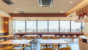A spacious dining area with high tables and chairs, large windows with a city view, and a wooden accent wall. - Hotel Monday Premium Ueno Okachimachi (Tokyo)