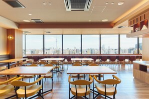 A spacious dining area with high tables and chairs, large windows with a city view, and a wooden accent wall. - Hotel Monday Premium Ueno Okachimachi (Tokyo)