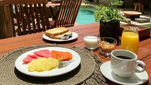 Een ontbijt met een fruitschaal, toast en een kop koffie op een houten tafel, met uitzicht op een zwembad en een gebouw met rieten dak.