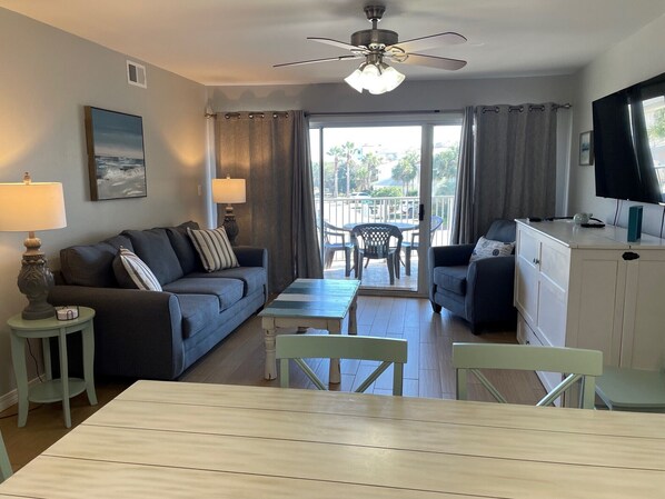 A modern living room with a sofa, a coffee table, and a dining table. There is a ceiling fan, a TV, and a window with curtains.