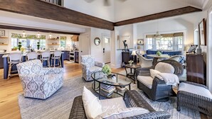 A spacious living area with a high ceiling, a dining area with a kitchen in the background, and a seating area with wicker furniture and a glass coffee table.