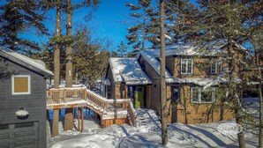 A cabin with a wooden deck and a green door, surrounded by snow-covered ground and tall trees.