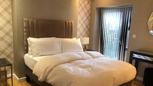 A modern hotel room with a large bed, a nightstand with a lamp, a window with curtains, and a small table. - 15% Off 4 Night Stays | Boutique 1 Bed Apartment (Cardiff)
