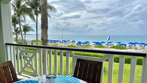 A balcony with a table, chairs, and a view of the beach with umbrellas. - Spectacular Beachfront Condo, Ocean Views of Grace Bay, Suite #123 (Providenciales)