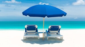 Two beach chairs under a blue umbrella on a white sandy beach with clear turquoise water and a clear sky. - Spectacular Beachfront Condo, Ocean Views of Grace Bay, Suite #123 (Providenciales)