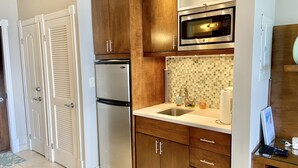 A compact kitchen with wooden cabinets, a stainless steel refrigerator, a microwave, and a tiled backsplash. - Spectacular Beachfront Condo, Ocean Views of Grace Bay, Suite #123 (Providenciales)
