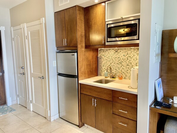 A compact kitchen with wooden cabinets, a stainless steel refrigerator, a microwave, and a tiled backsplash. - Spectacular Beachfront Condo, Ocean Views of Grace Bay, Suite #123 (Providenciales)