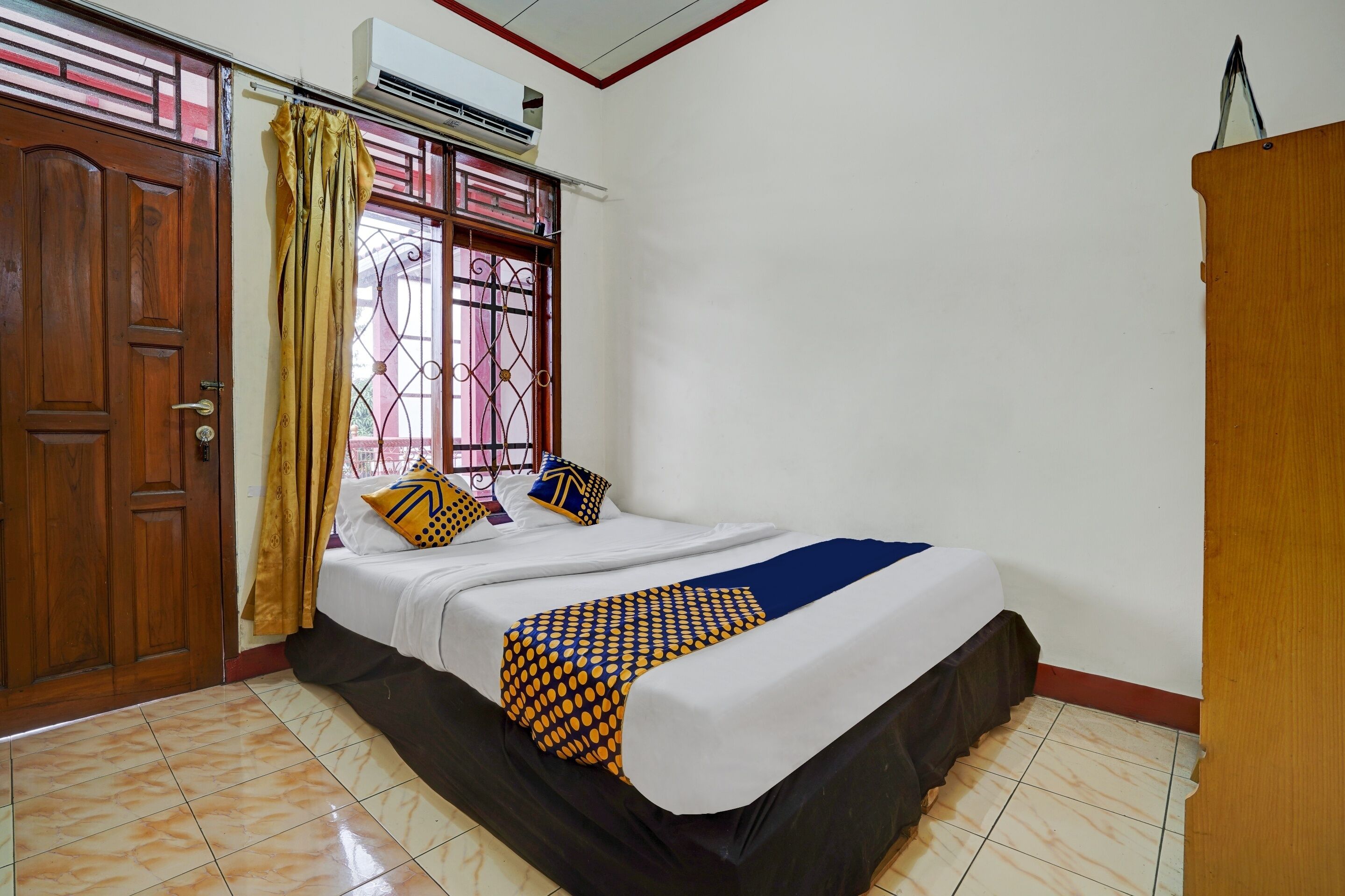 A bedroom with a bed, curtains, a window with a metal grill, a wooden door, and a wardrobe.