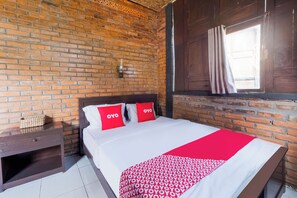 Habitación de hotel con pared de ladrillo, cabecero de madera, cama con ropa blanca y almohadas rojas, mesita de noche con lámpara y ventana con cortinas.